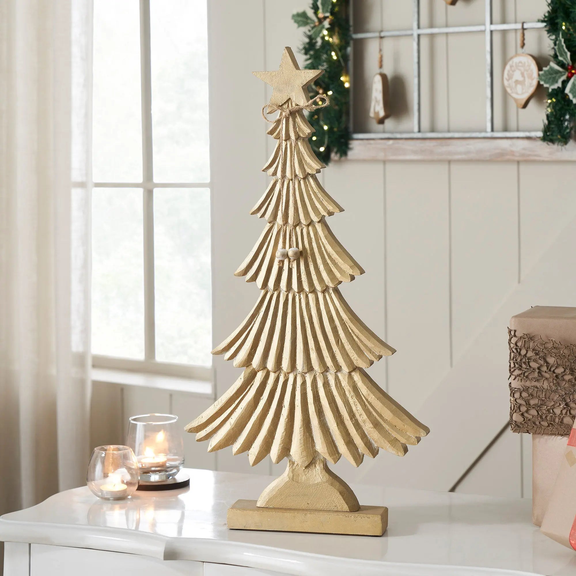 Decorative gold Christmas tree on a table with candles and a window in the background.