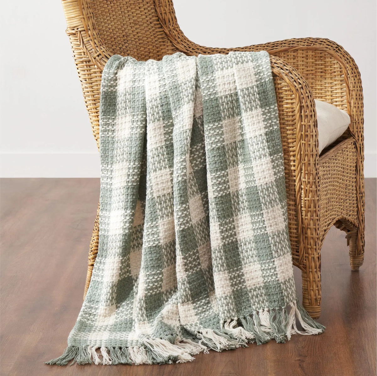 A woven throw blanket with a sage green and cream plaid pattern and fringed edges, draped over a wicker armchair on a wooden floor.