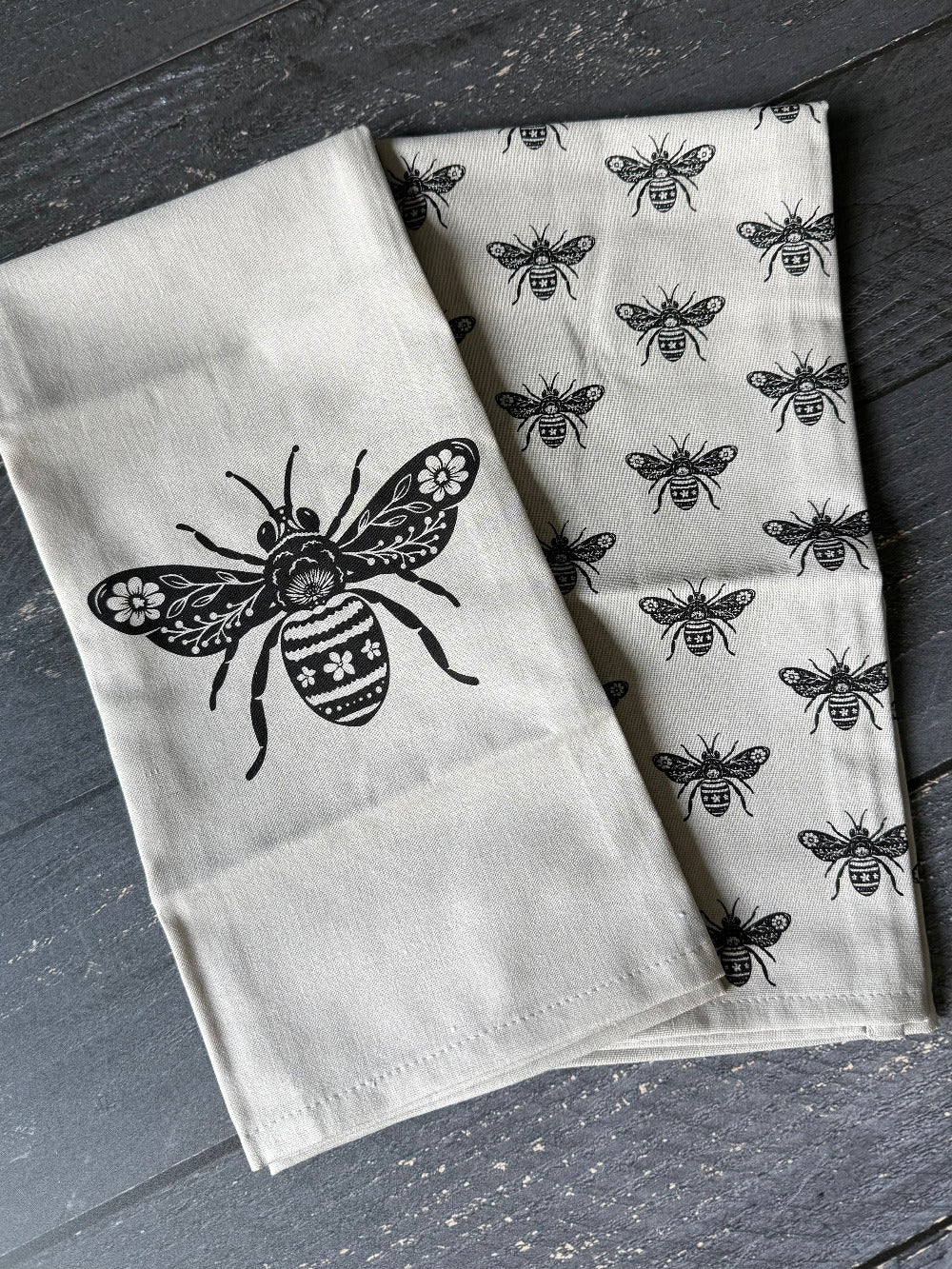 Two white towels with black bee patterns on a dark wooden surface