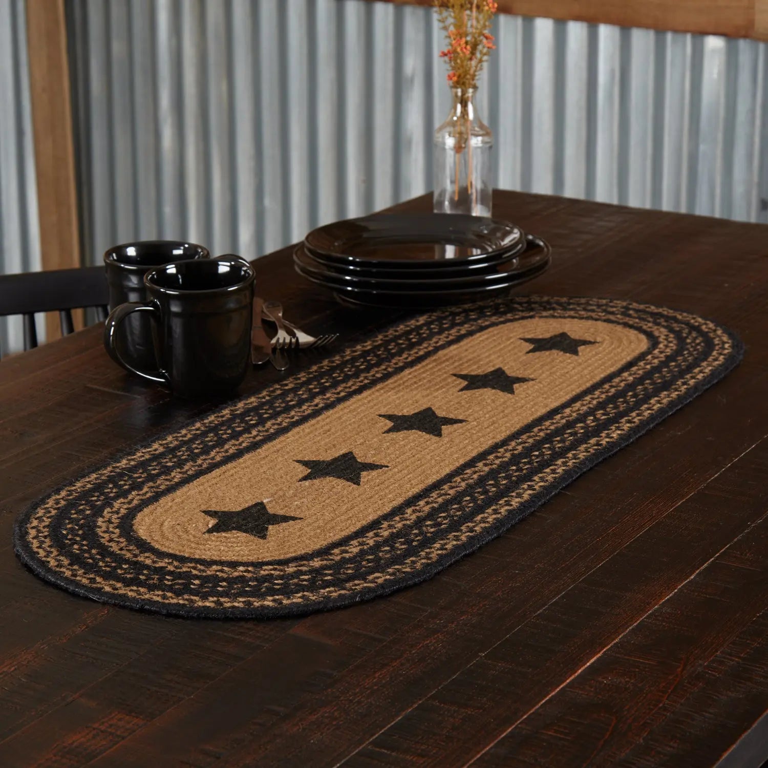An oval braided table runner with four black stars, styled on a dark wood dining table next to black mugs and plates.