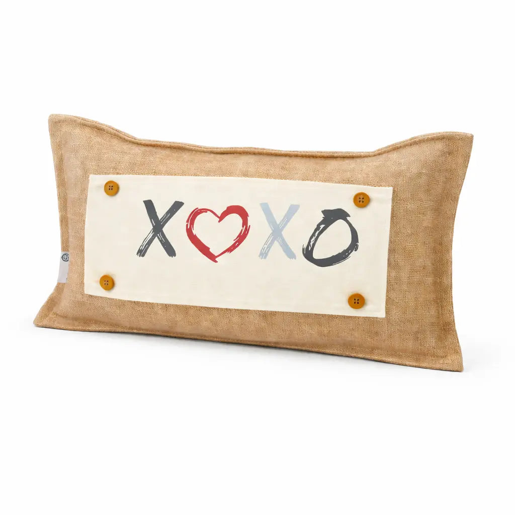 A rectangular beige linen throw pillow featuring a central cream fabric patch with the letters 'X', a red heart, 'X', and 'O' in black and light blue, accented by four small orange buttons in the corners.