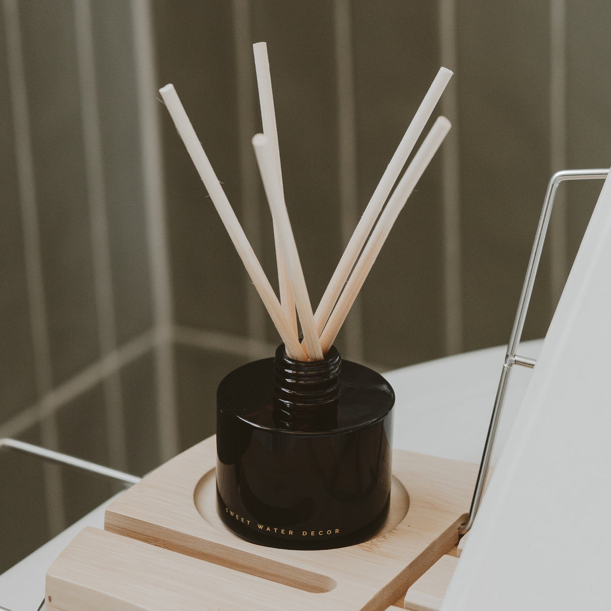 A reed diffuser with a glossy black bottle and light natural wood reeds, displayed on a light wood stand against a soft gray background.