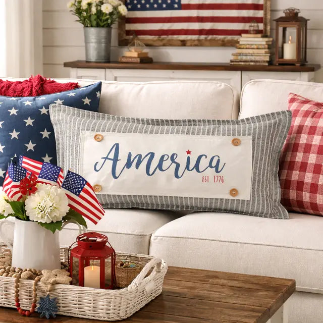 A rectangular decorative throw pillow with a gray and white striped border and a white center panel reading 'America Est. 1776' in blue script, styled on a white sofa in a patriotic living room setting.