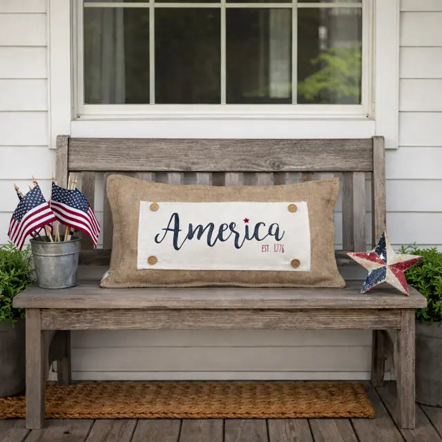A rustic burlap throw pillow with 'America Est. 1776' printed on the front, styled on a weathered wooden bench on a front porch with American flags and a red, white, and blue star decoration.