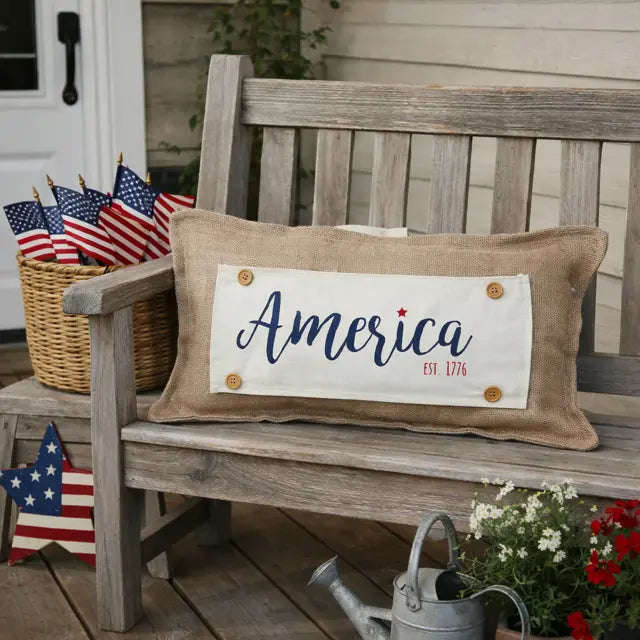 A rustic burlap throw pillow with a white patch reading 'America Est. 1776' sits on a weathered wooden porch bench.