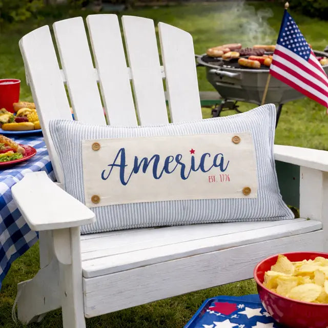 A white wooden Adirondack chair outdoors on grass, styled with a patriotic throw pillow that reads 'America Est. 1776' in blue script on a cream panel.