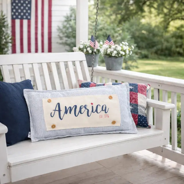 A rectangular decorative throw pillow with a patriotic design sits on a white porch swing. The pillow features a cream center panel with the text 'America Est 1776' in navy blue script and red block lettering, bordered by a light blue and white striped fabric.