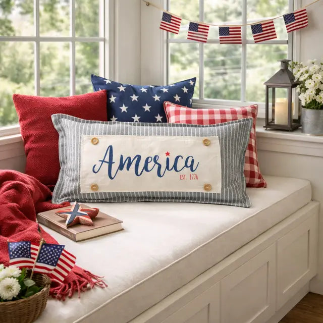 A patriotic-themed home decor scene featuring a central rectangular throw pillow with 'America Est. 1776' embroidered in blue script on a cream background with gray stripes.