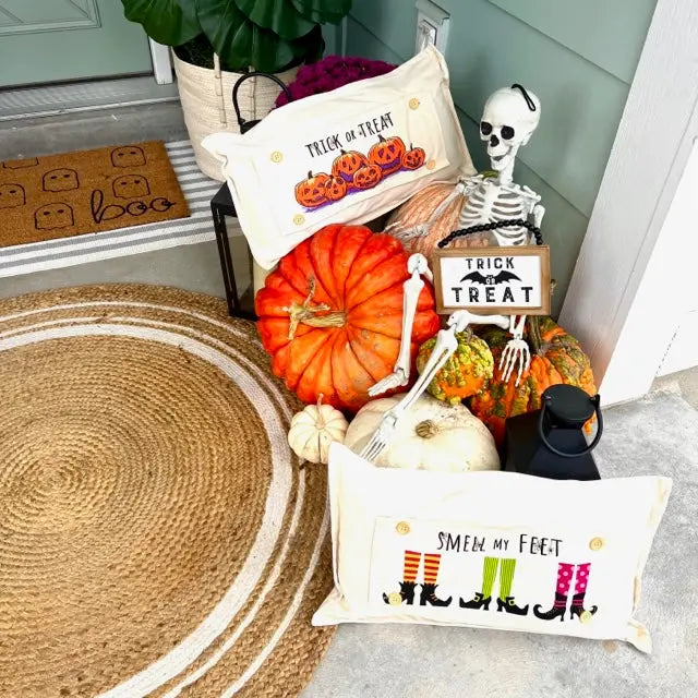 A styled Halloween entryway display featuring two cream decorative pillows with autumn-themed prints, surrounded by pumpkins, a skeleton decoration, a small sign reading 'Trick or Treat,' and other seasonal decor on a porch step.
