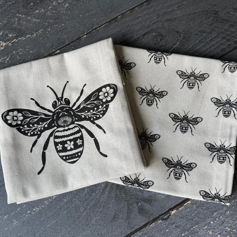 Two folded cream-colored linen napkins with black printed bee designs, displayed on a dark gray wooden surface.