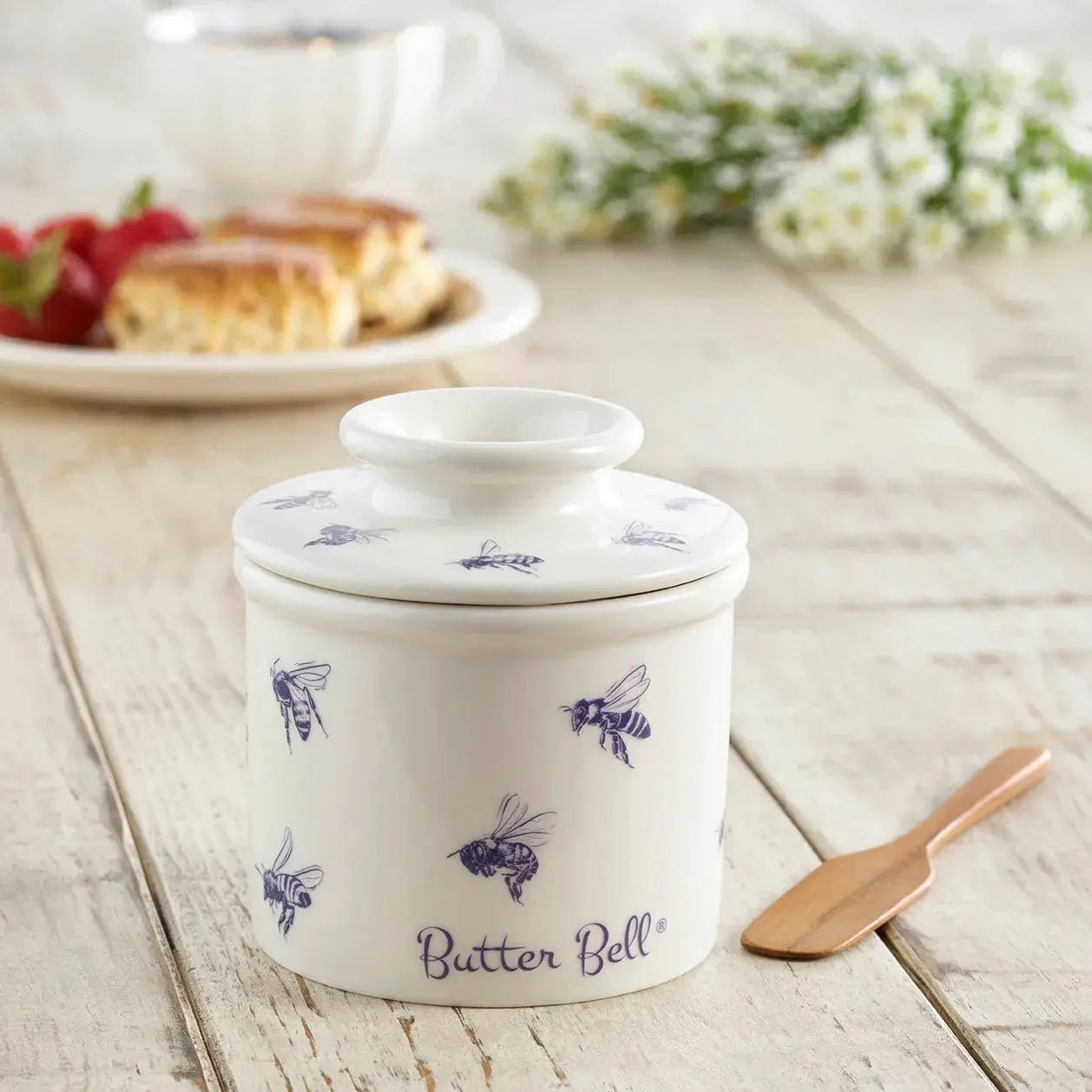 White ceramic butter bell with blue bee print, farmhouse kitchen decor, wooden table with scones and flowers