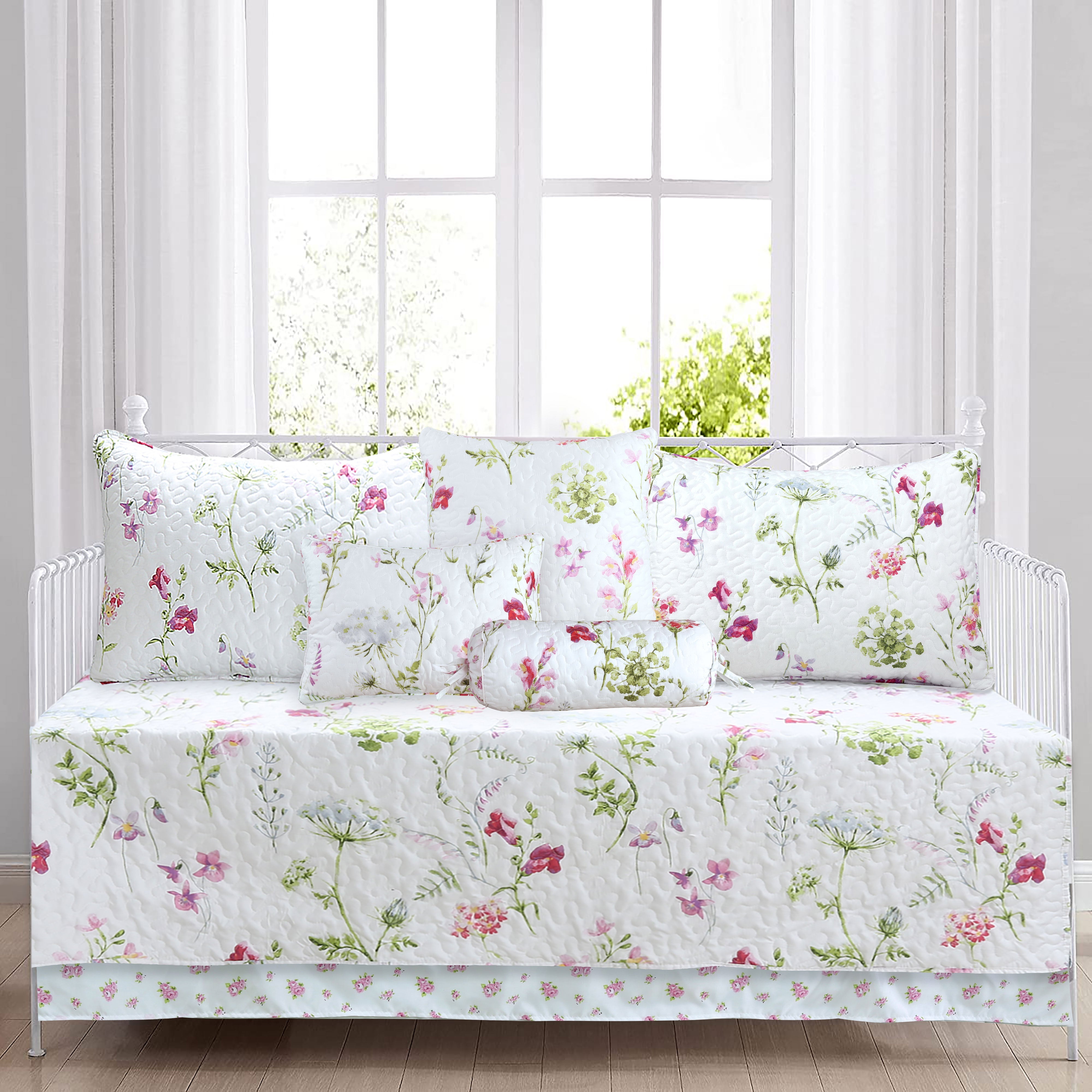 A white quilted daybed cover set with a pink and green floral print, including a matching bed skirt, comforter, and three pillows, displayed on a white metal daybed in front of a bright window.