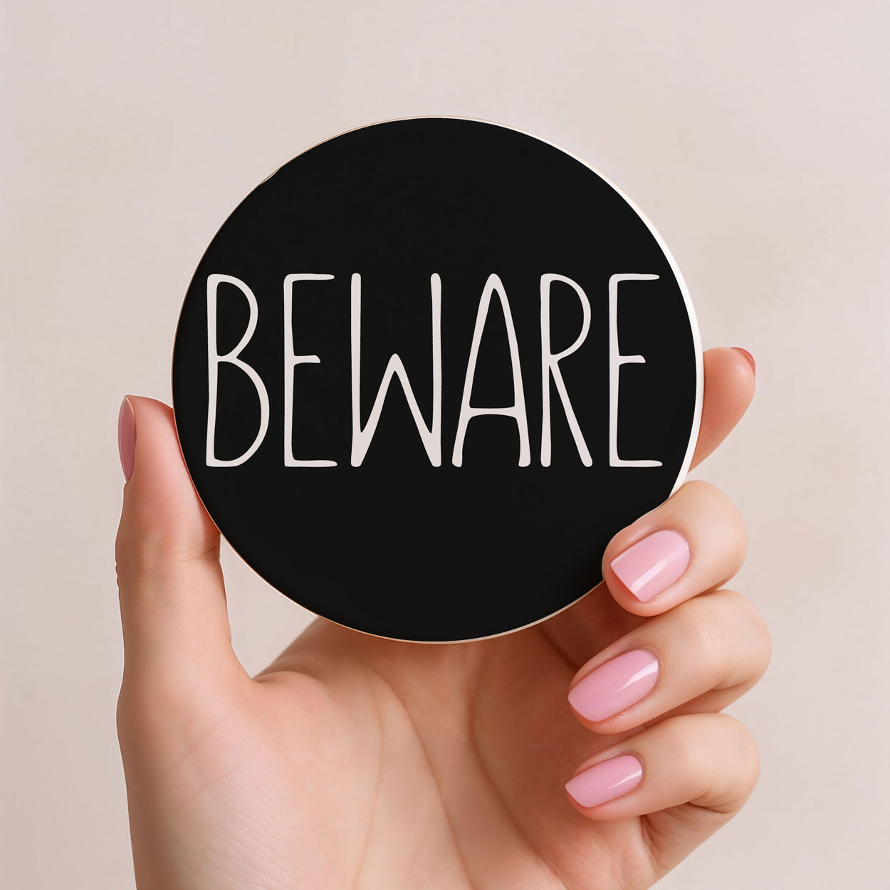 Hand holding a round black sign with 'BEWARE' text on a beige background
