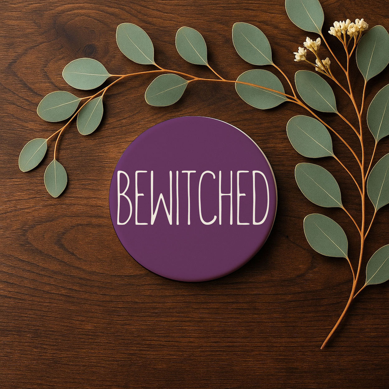 Purple circular sign with 'BEWITCHED' text on a wooden surface with leaves and branches.