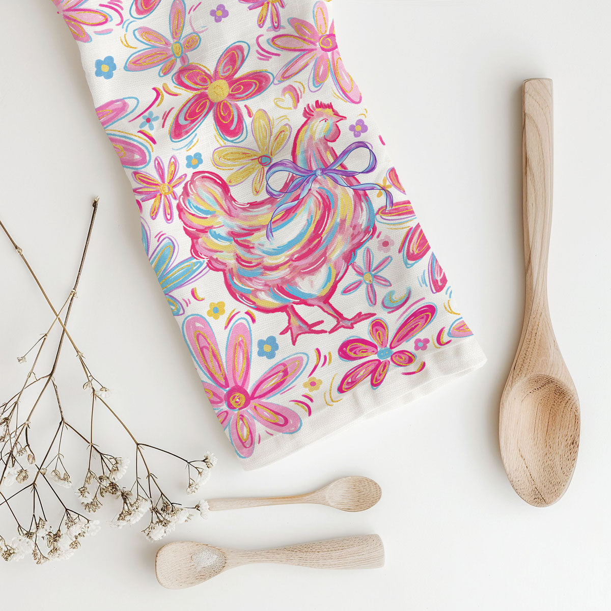 Colorful towel with rooster design on a white surface with wooden spoons and dried flowers.
