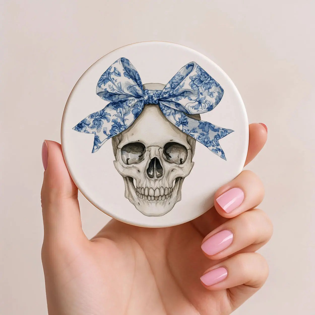 Hand holding a round object with a skull design and blue bow on a beige background