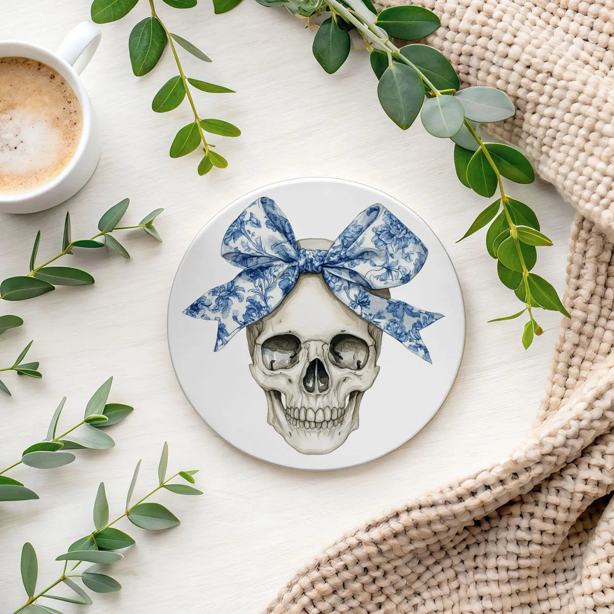 Coaster with skull design and blue bow surrounded by green leaves and a cup of coffee.