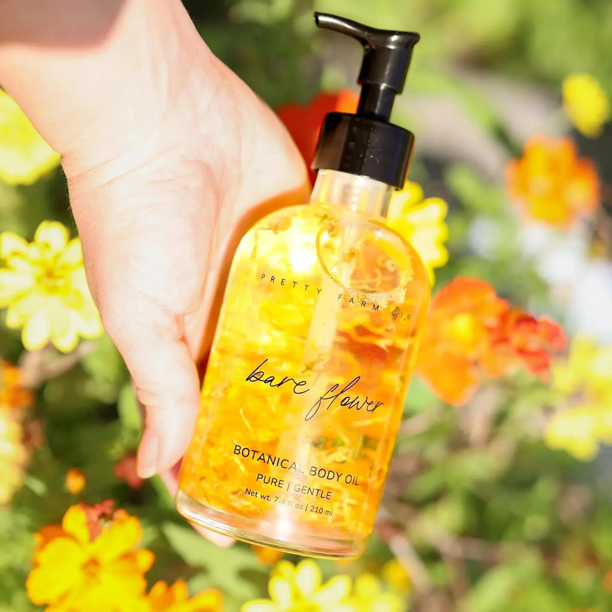 Hand holding botanical body oil with flower petals, labeled 'bare flower' against garden blooms