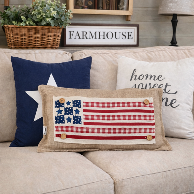 A rectangular beige throw pillow with a rustic American flag design made from red-and-white gingham fabric, blue fabric stars, and small wooden buttons, styled on a beige sofa with navy and cream decorative pillows in a living room setting.