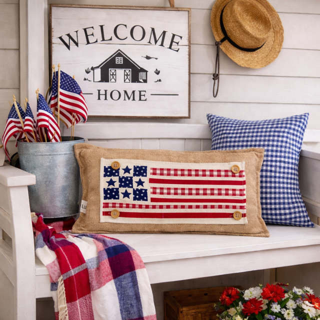 Decorative American flag pillow on a bench with 'Welcome Home' sign and American flags in the background.