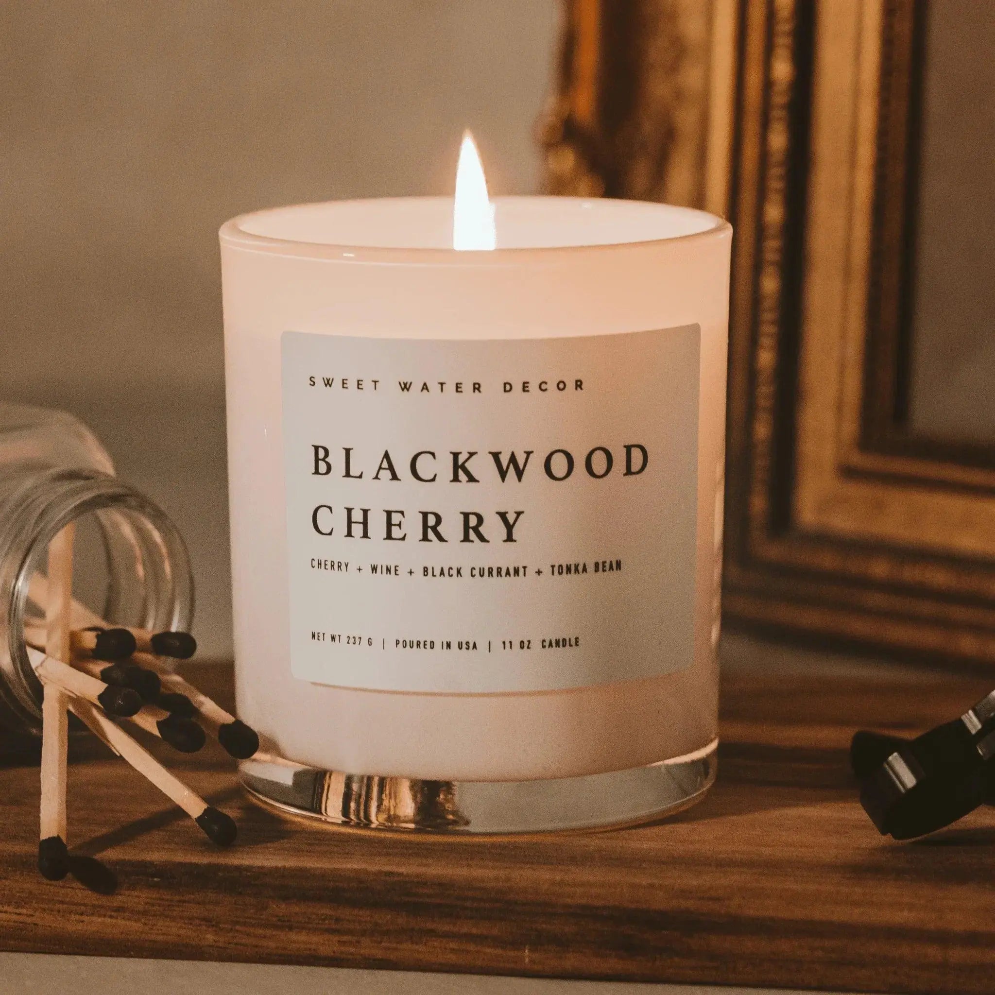 Blackwood Cherry scented candle lit on wooden surface, cozy farmhouse decor with matches and gold frame