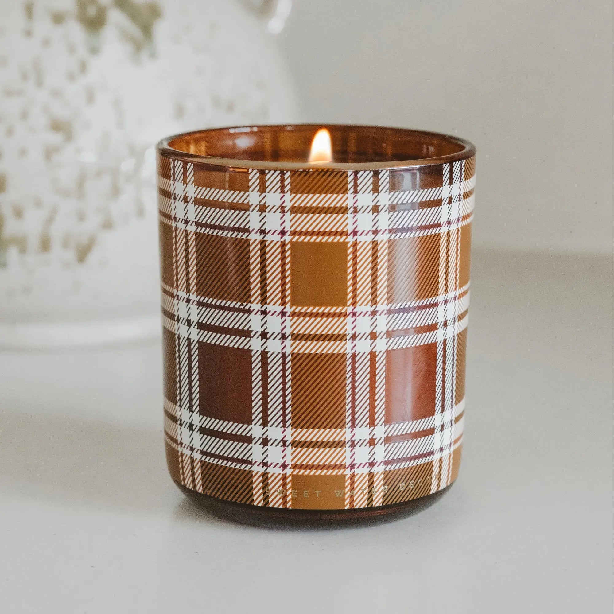 Candle in a brown glass container with plaid pattern on a white surface.