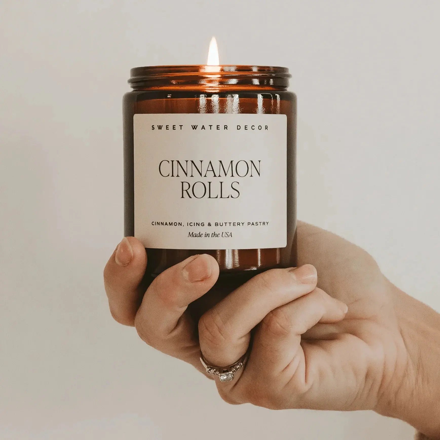 Hand holding a candle labeled 'Cinnamon Rolls' by Sweet Water Decor against a neutral background