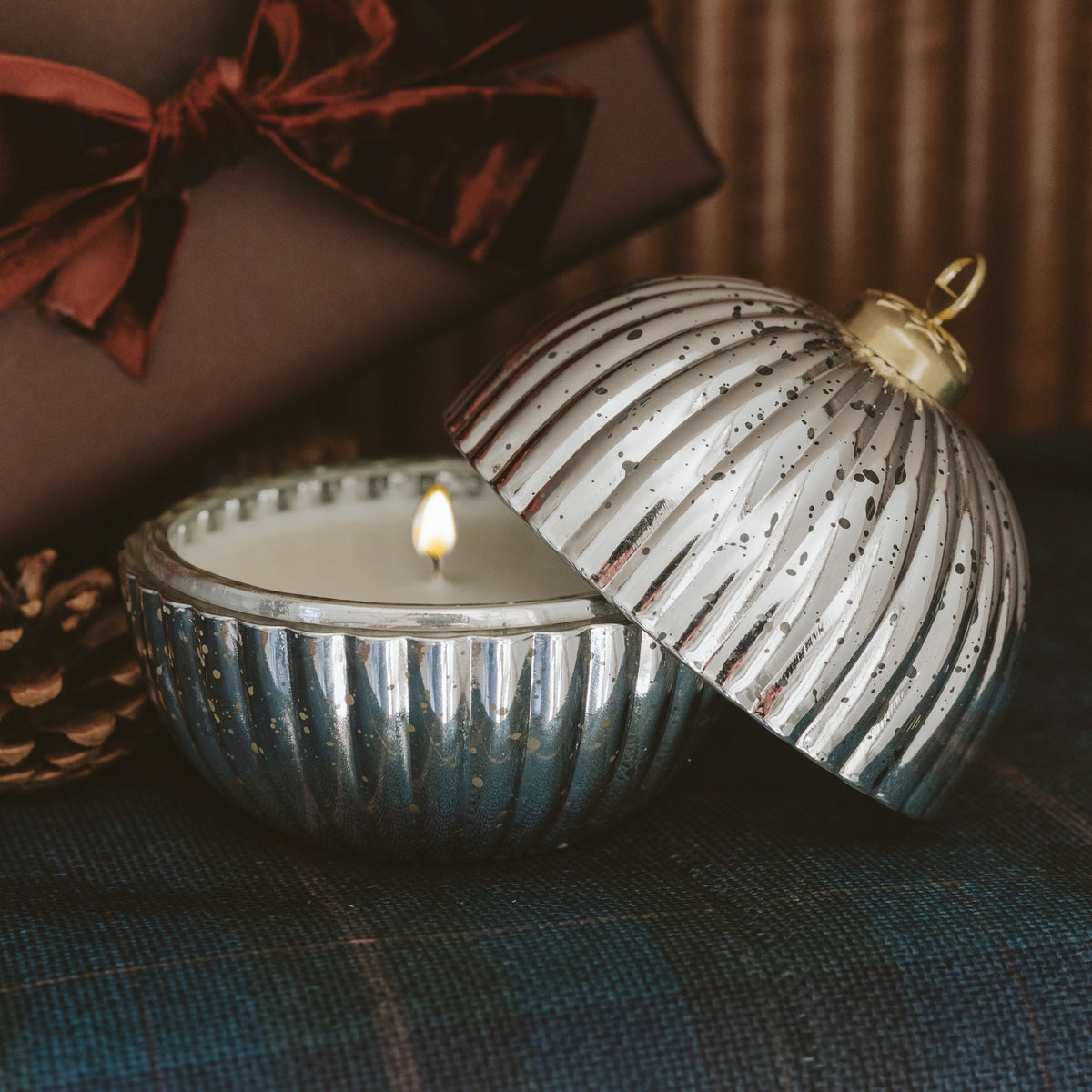 A silver ribbed glass jar candle with its lid propped open, showing a lit white candle inside. The candle is styled on a dark blue plaid fabric with a brown gift box and ribbon in the background.
