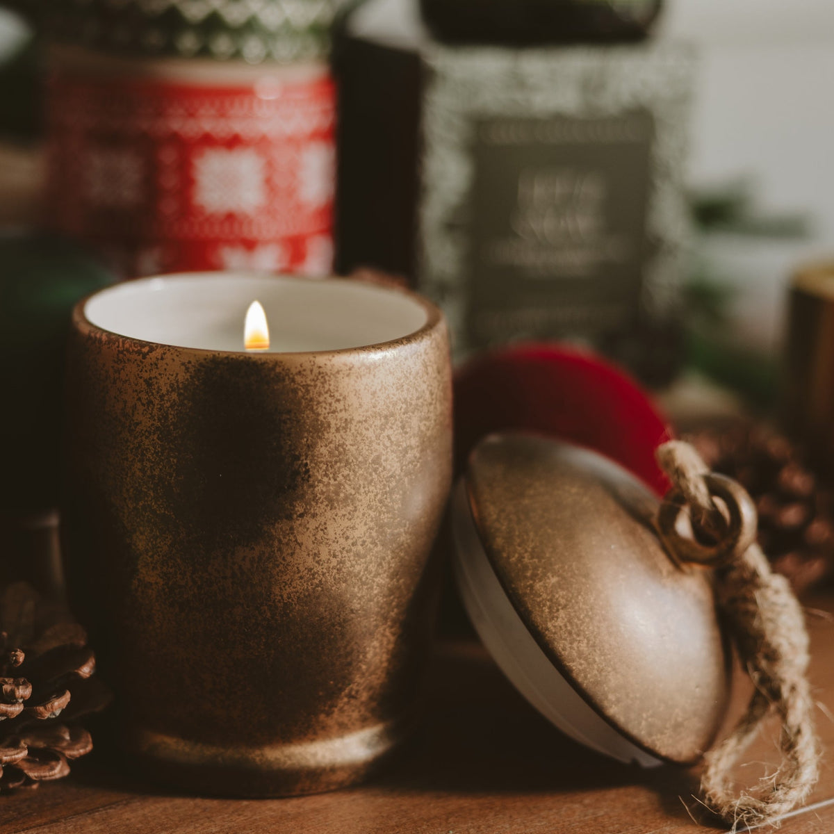 A lit candle in a textured metallic bronze ceramic holder sits on a wooden surface next to its matching lid, with blurred holiday decorations in the background.
