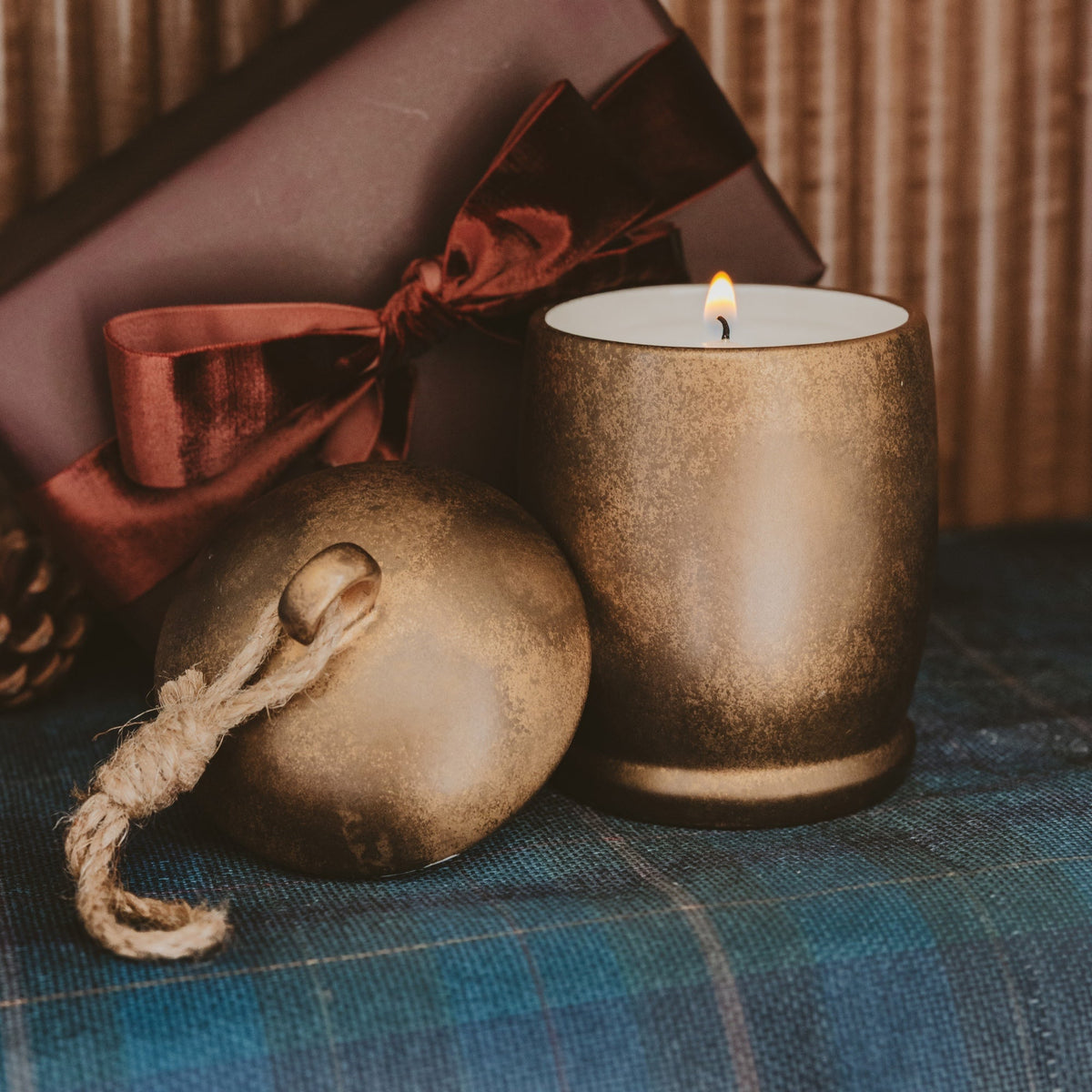 A lit candle in a textured metallic gold holder sits beside a matching gold ornament with a rope loop, with a dark gift box and ribbon in the background on a blue plaid surface.