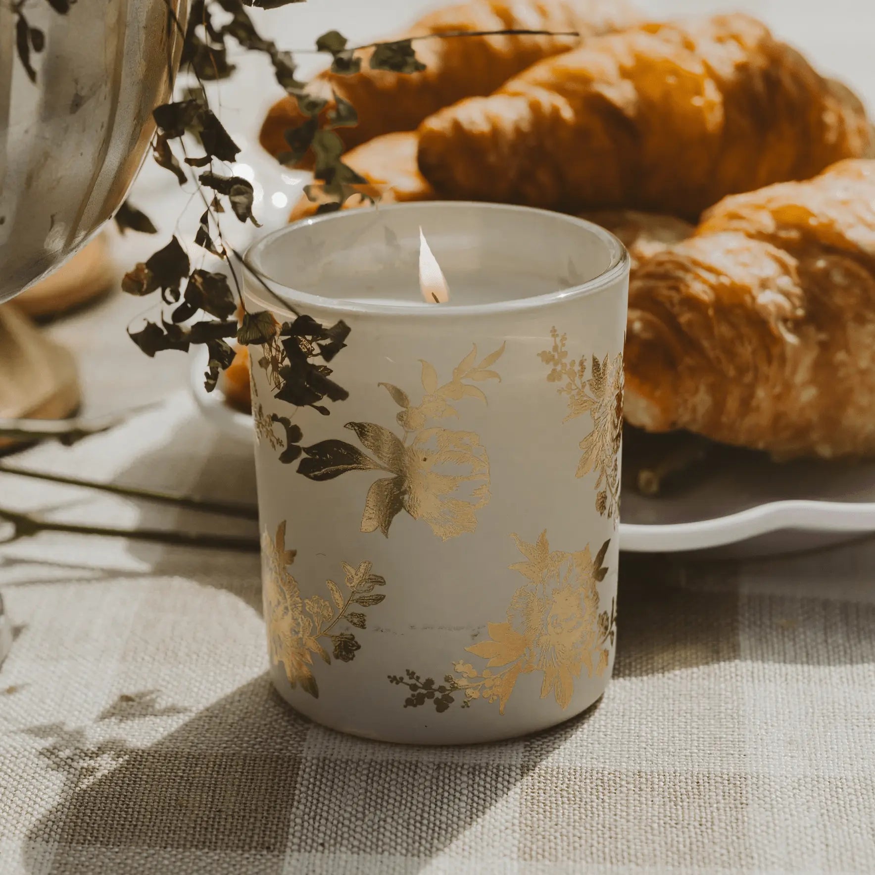Lit floral gold candle on checkered cloth with croissants, cozy farmhouse home decor