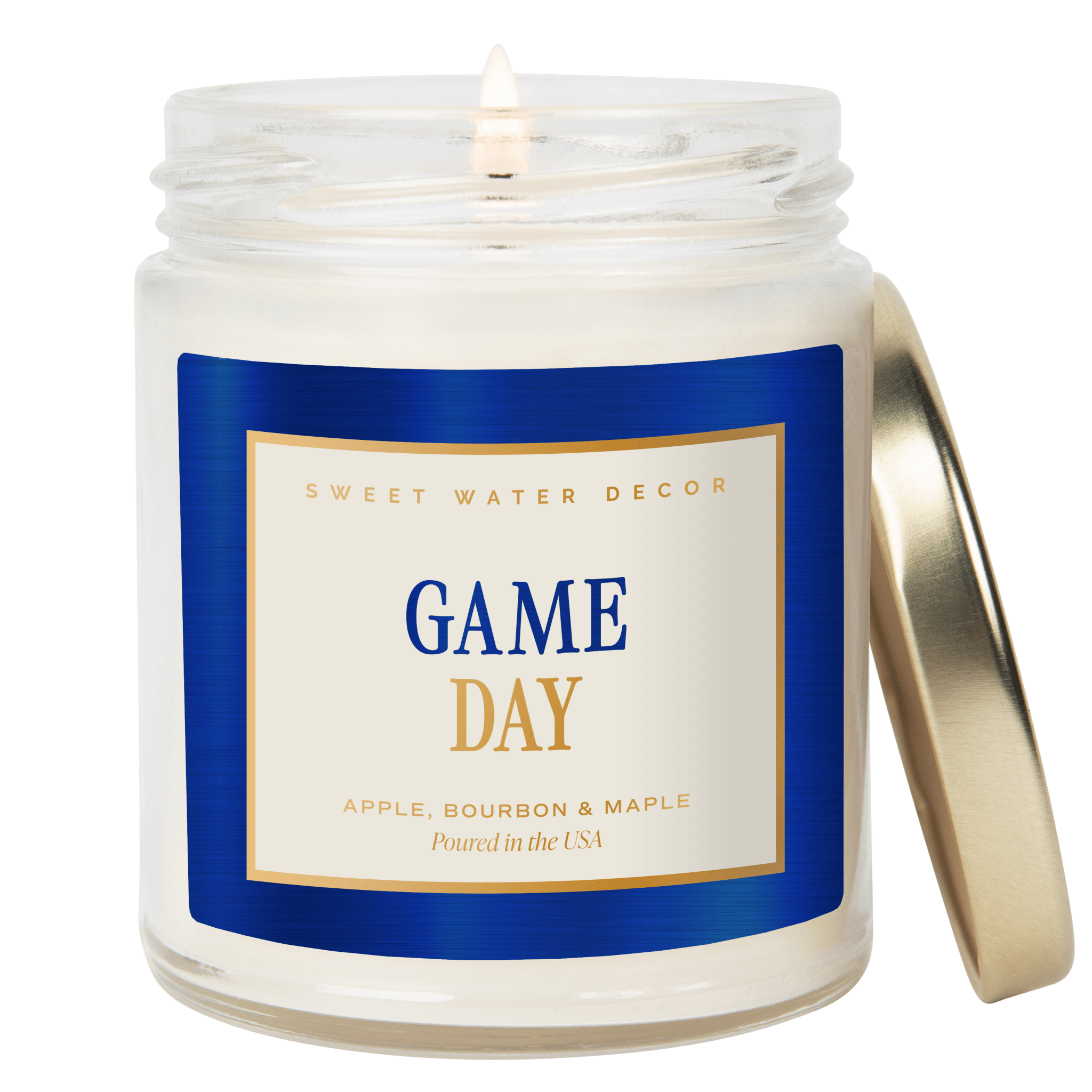 An open white jar candle with a lit wick, featuring a blue and cream label that reads 'GAME DAY' and 'SWEET WATER DECOR.' A metallic gold lid leans against the right side of the jar.