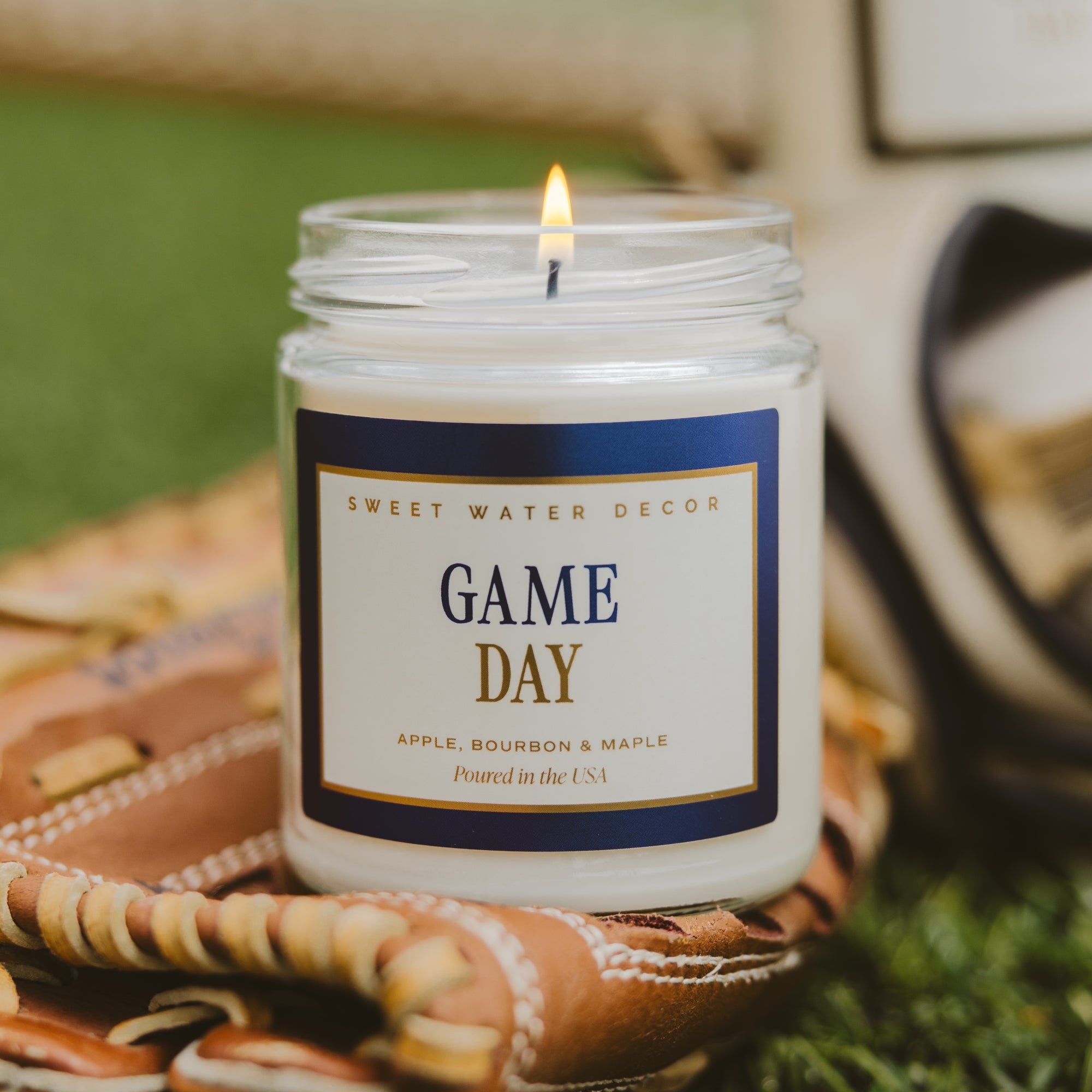 A lit white candle in a clear glass jar sits on a baseball glove outdoors. The front label reads “Sweet Water Decor” and “Game Day,” with apple, bourbon, and maple notes.
