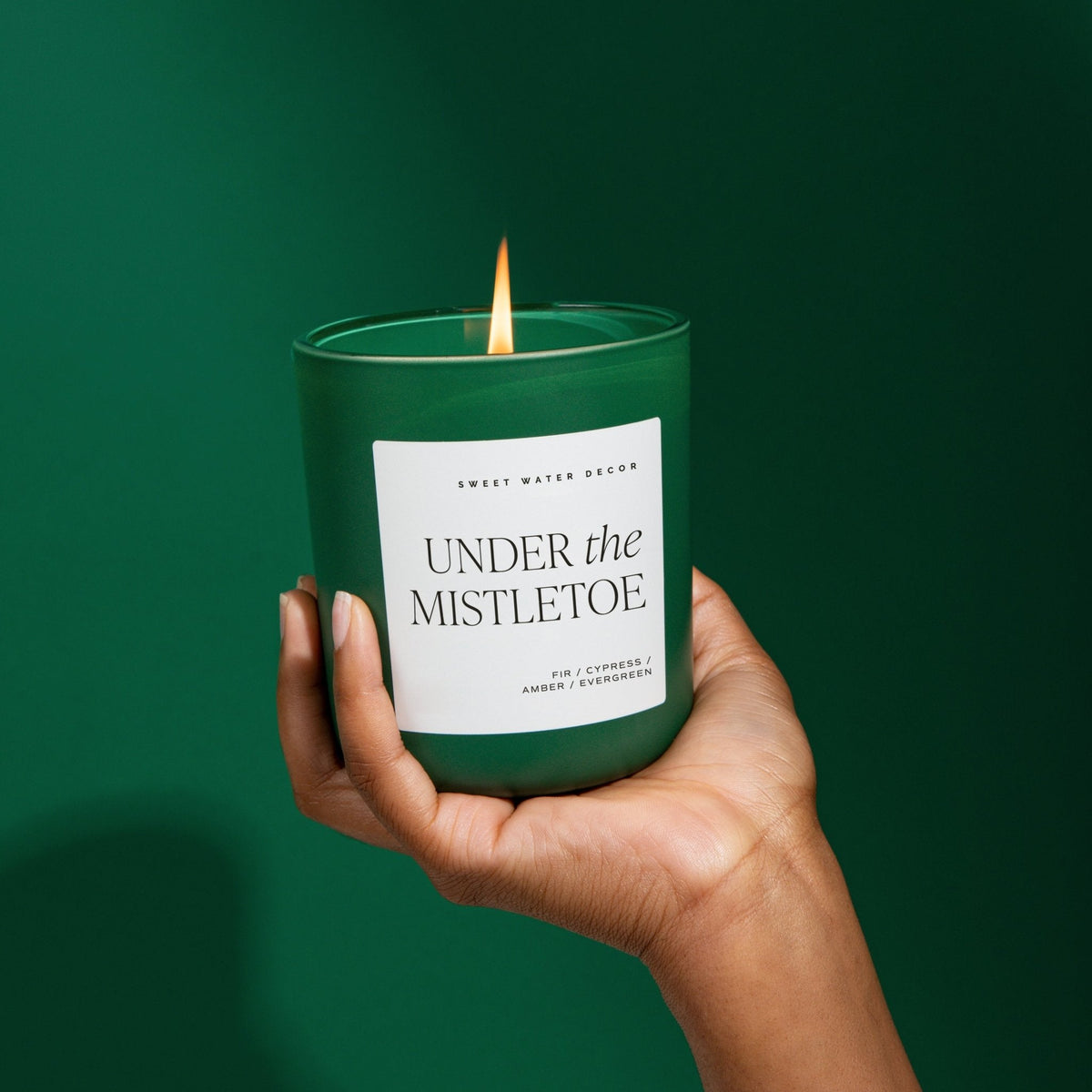 Hand holding a green candle labeled 'Under the Mistletoe' against a dark green background