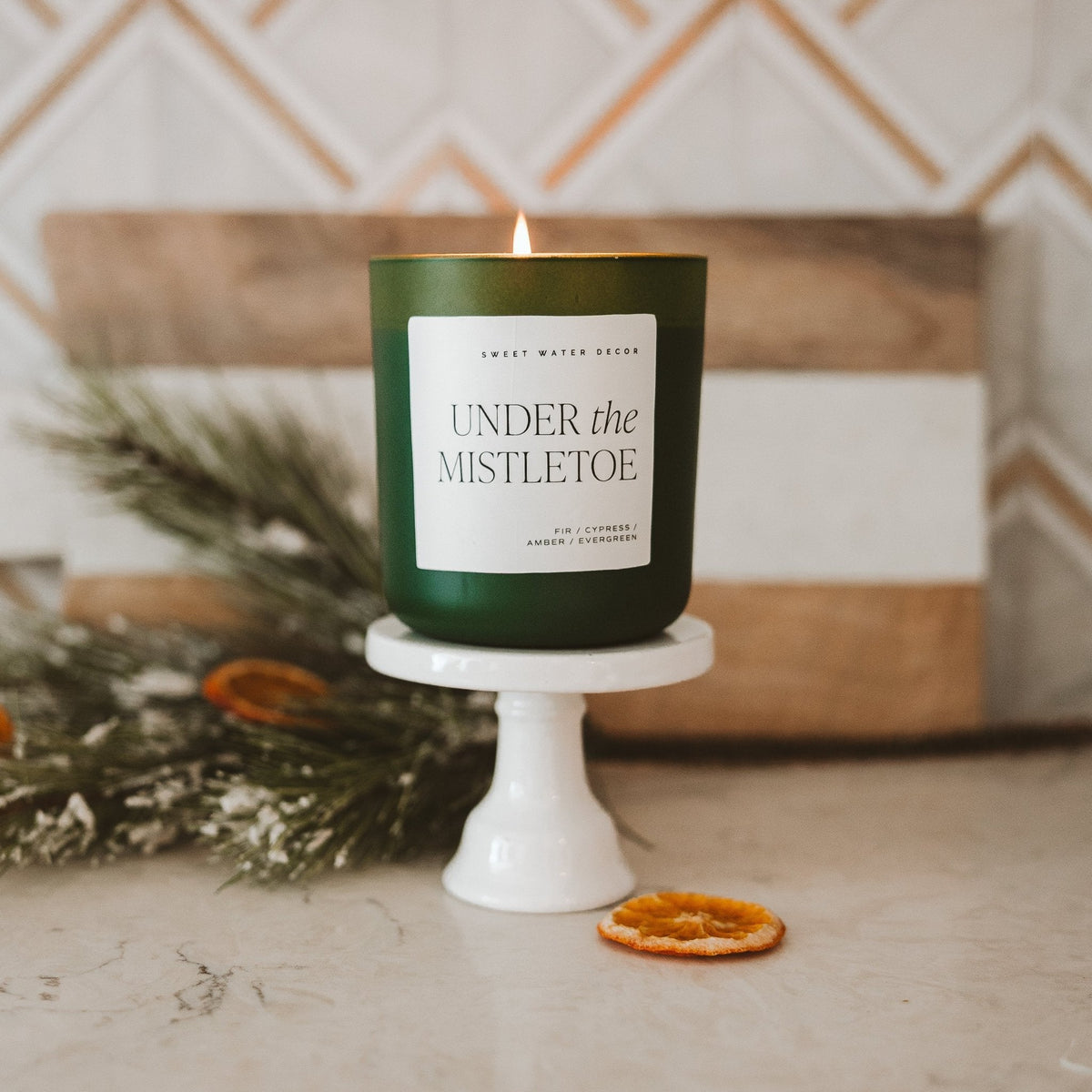 Green candle labeled 'Under the Mistletoe' on a white stand with decorative elements.