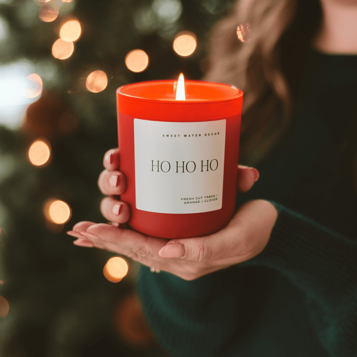 Person holding a red candle labeled 'HO HO HO' with Christmas lights in the background