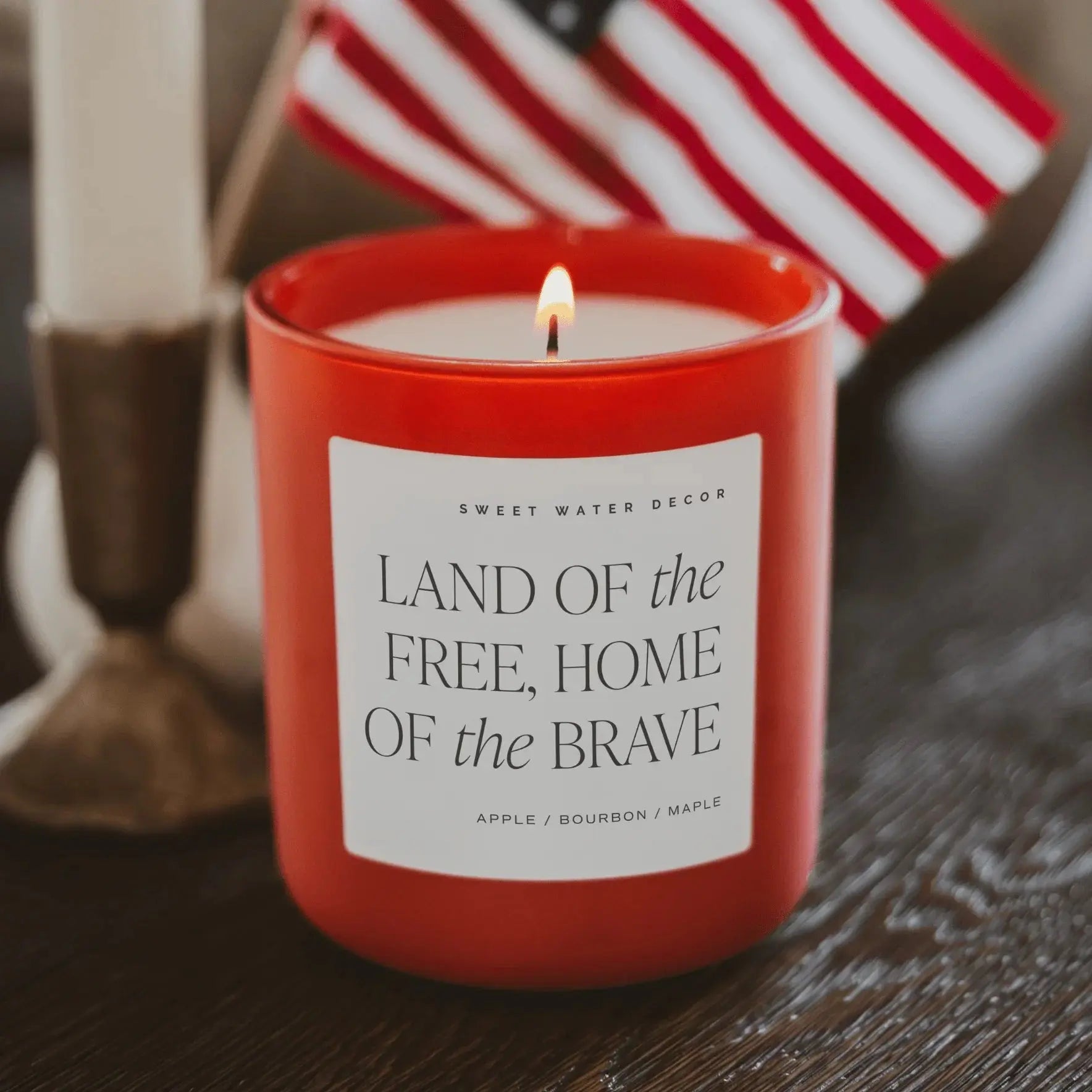 Red candle with a label on a wooden surface, featuring an American flag in the background.