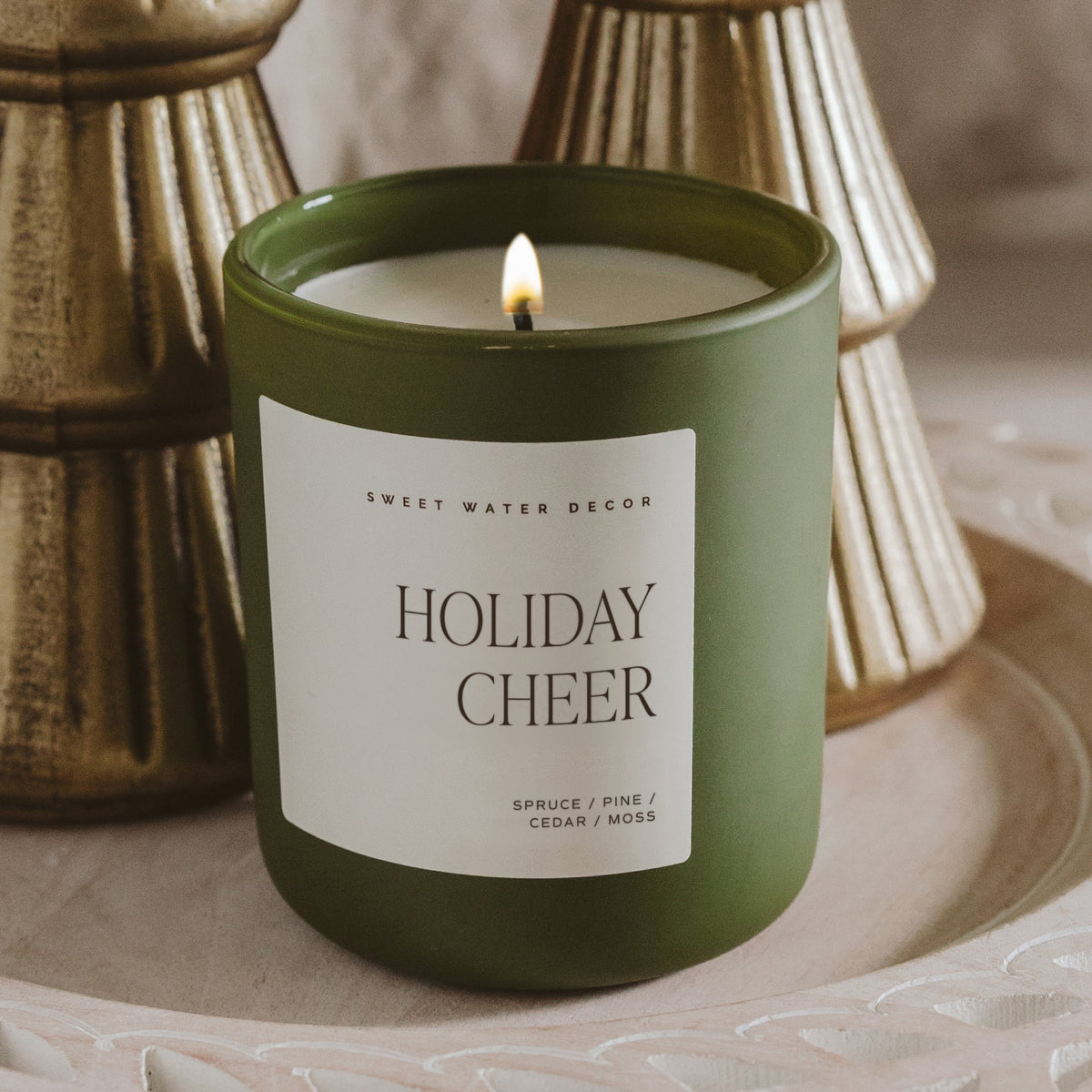 A green jar candle with a white label reading 'HOLIDAY CHEER' by Sweet Water Decor, lit and set on a decorative tray with blurred gold-toned vessels in the background.