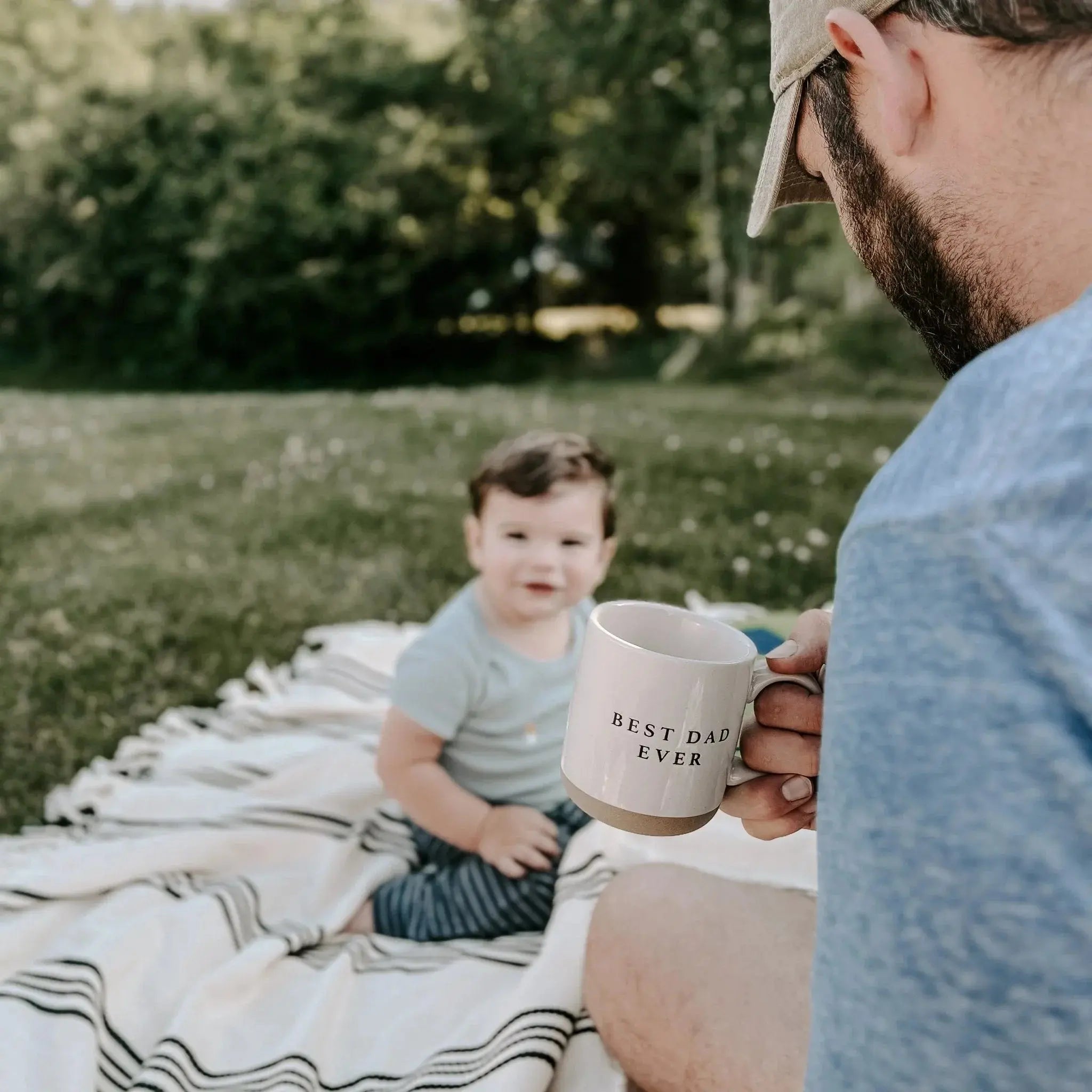 Dad holding 'Best Dad Ever' mug with baby on picnic blanket, cozy outdoor family moment