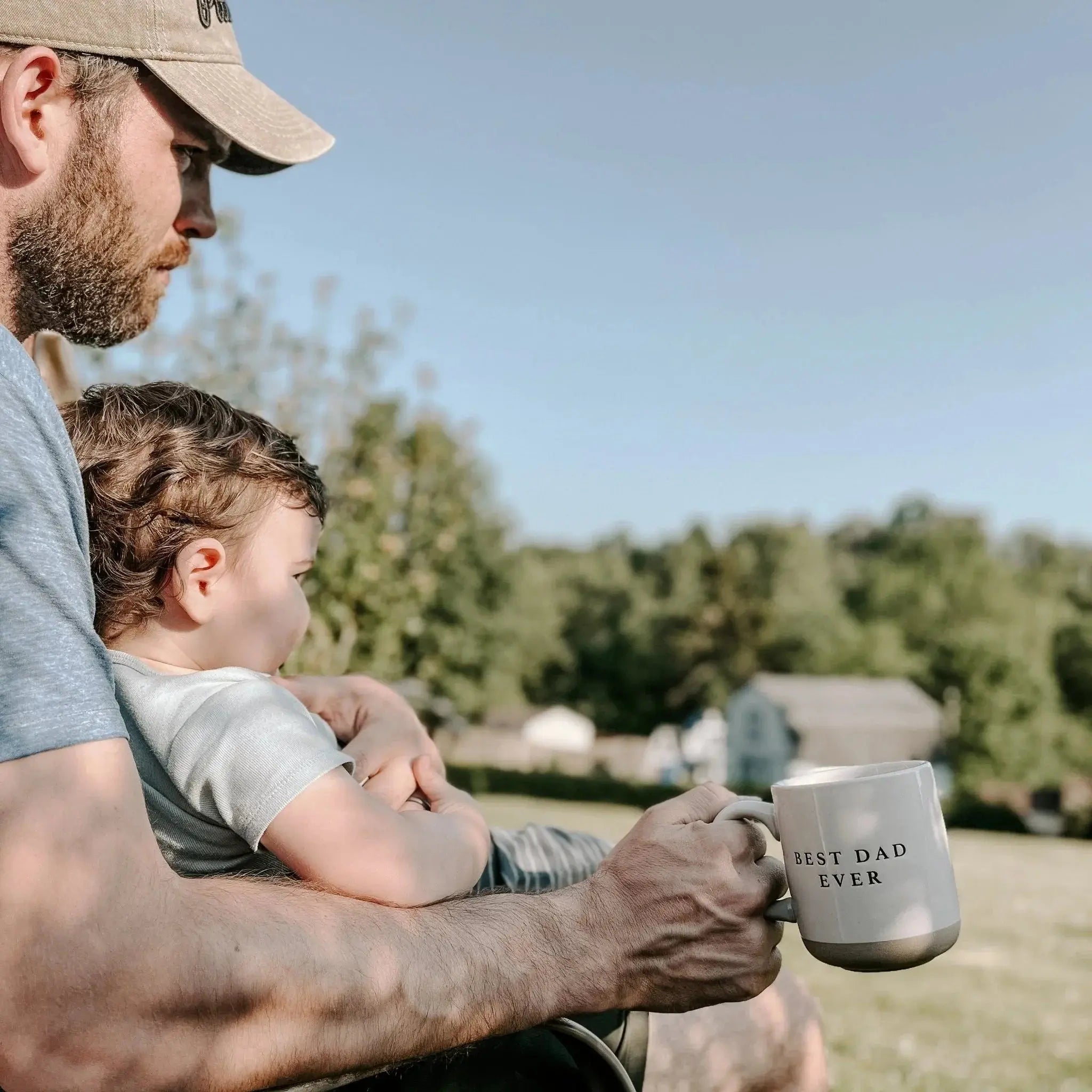 Dad holding child outdoors with 'Best Dad Ever' coffee mug, cozy family moment