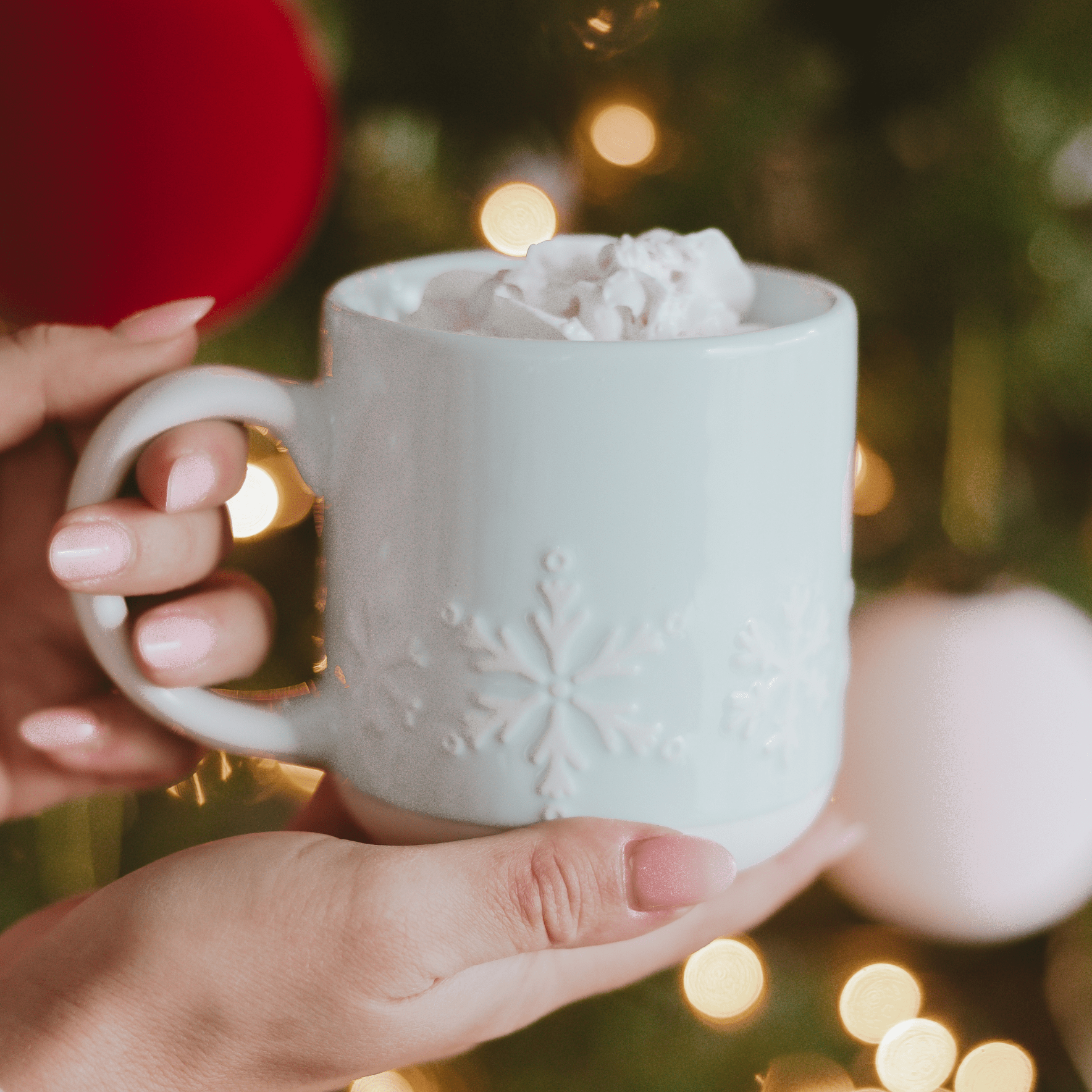 White mug with snowflake design held by a hand against a festive background with lights and ornaments.