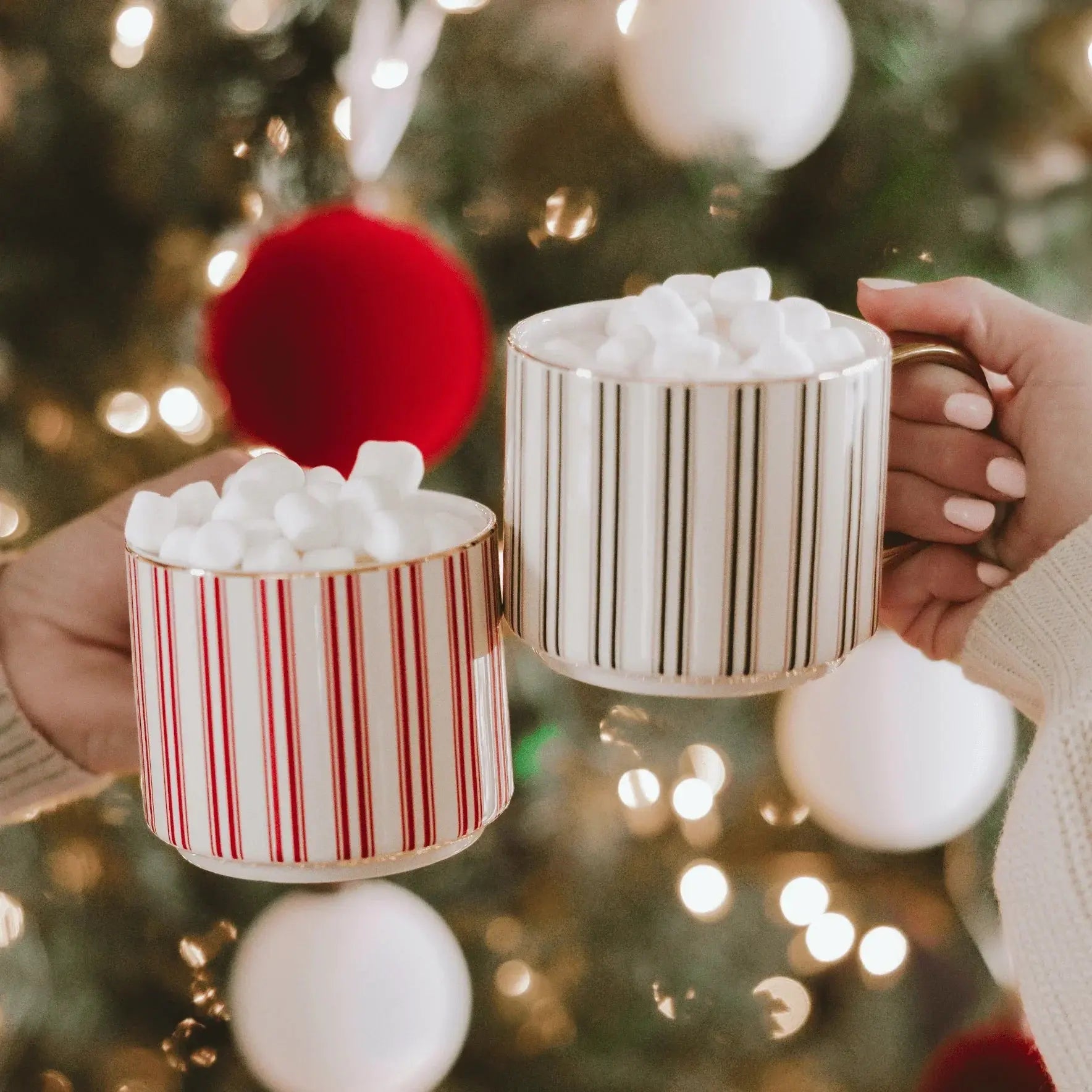 Cozy holiday mugs with marshmallows, striped designs, held near Christmas tree lights