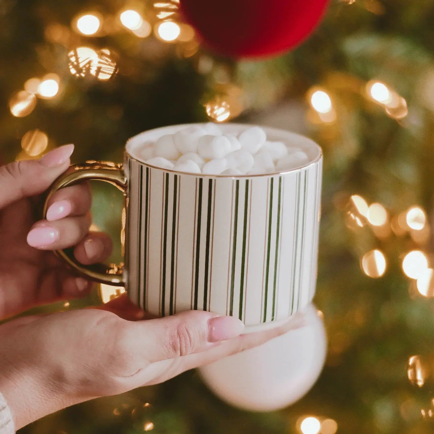 Cozy striped mug with marshmallows, held near glowing Christmas tree lights for festive farmhouse decor