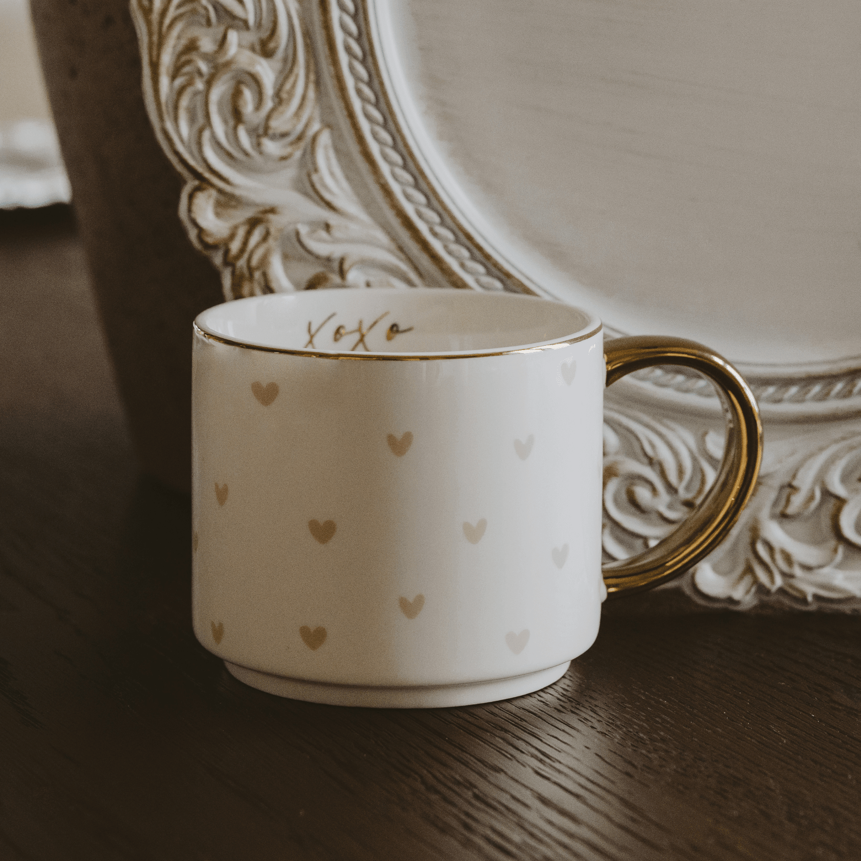 White mug with gold hearts and 'XOXO' text on a wooden surface with decorative plate in the background