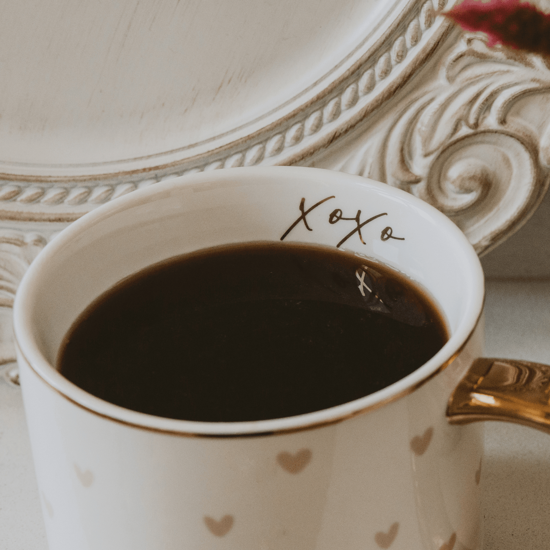 White mug with gold hearts and 'xoxo' text, filled with dark liquid, on a decorative surface.