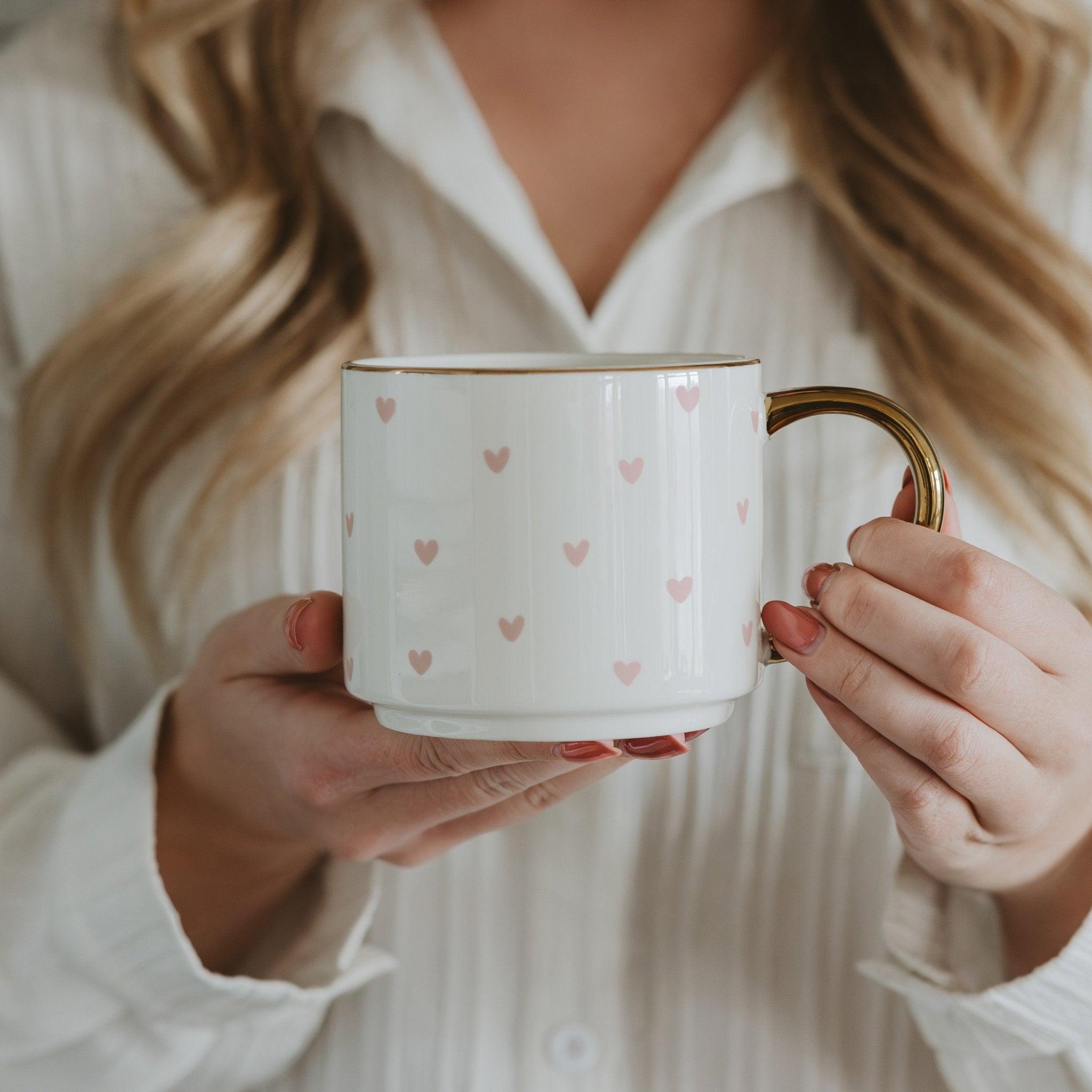 Person holding a white mug with pink heart patterns and a gold handle.