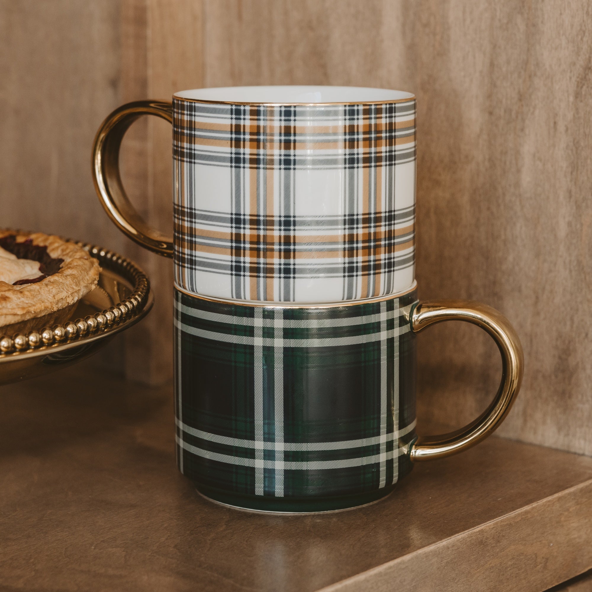 Two stacked ceramic mugs with gold-tone handles, featuring plaid patterns. The top mug has a white, brown, and navy plaid design, while the bottom mug has a dark green, white, and gray plaid design.