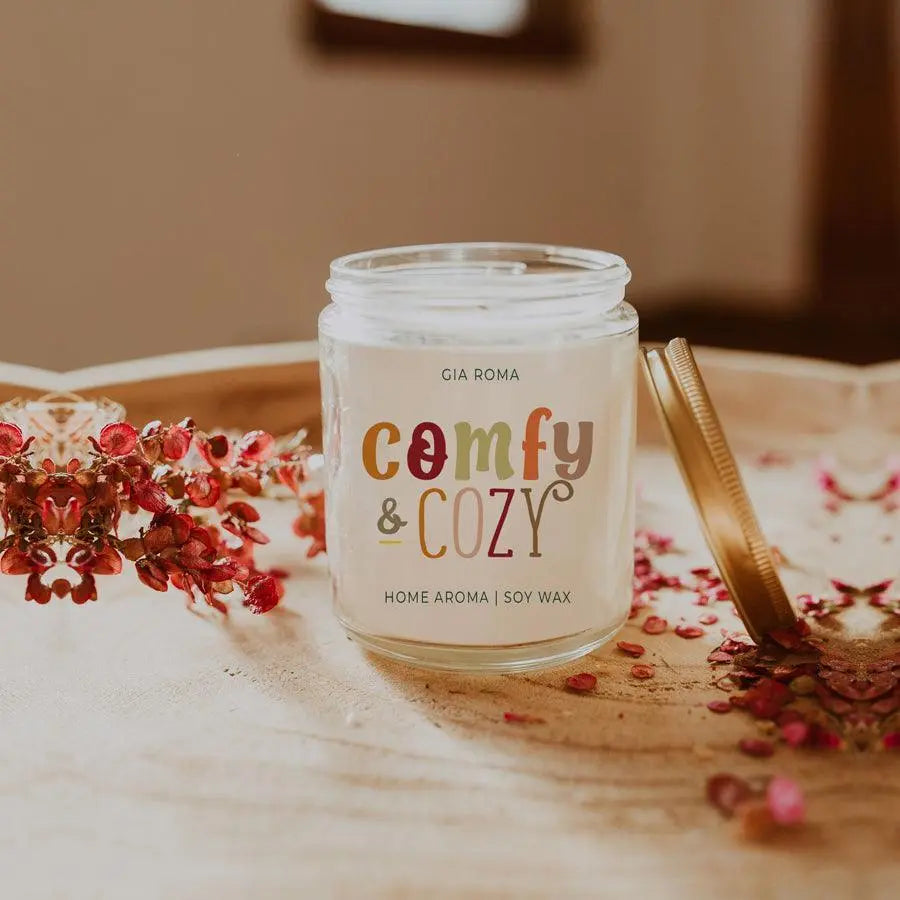 Gia Roma 'Comfy & Cozy' candle on a decorative surface with flowers and a mirror.