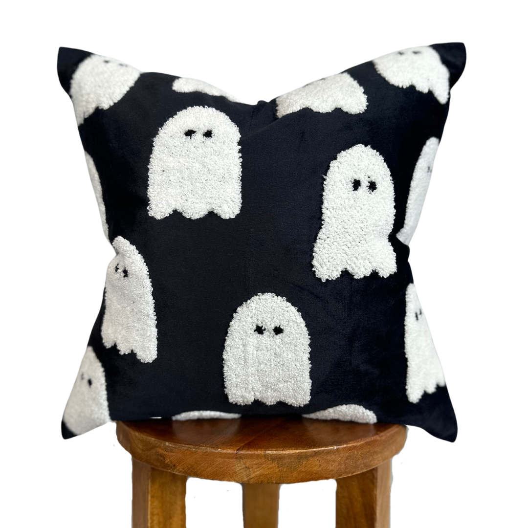 A square black throw pillow with a white textured ghost pattern, displayed on a wooden stool against a plain white background.