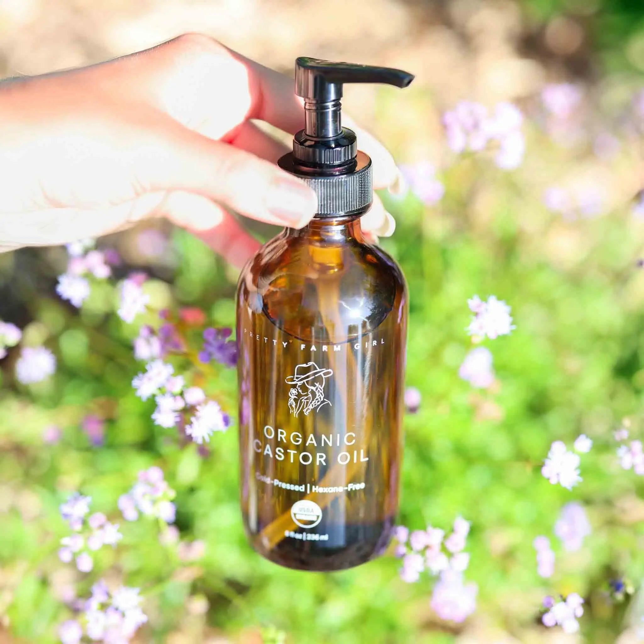 Organic castor oil in amber glass bottle with pump, held outdoors among wildflowers, natural bath essential